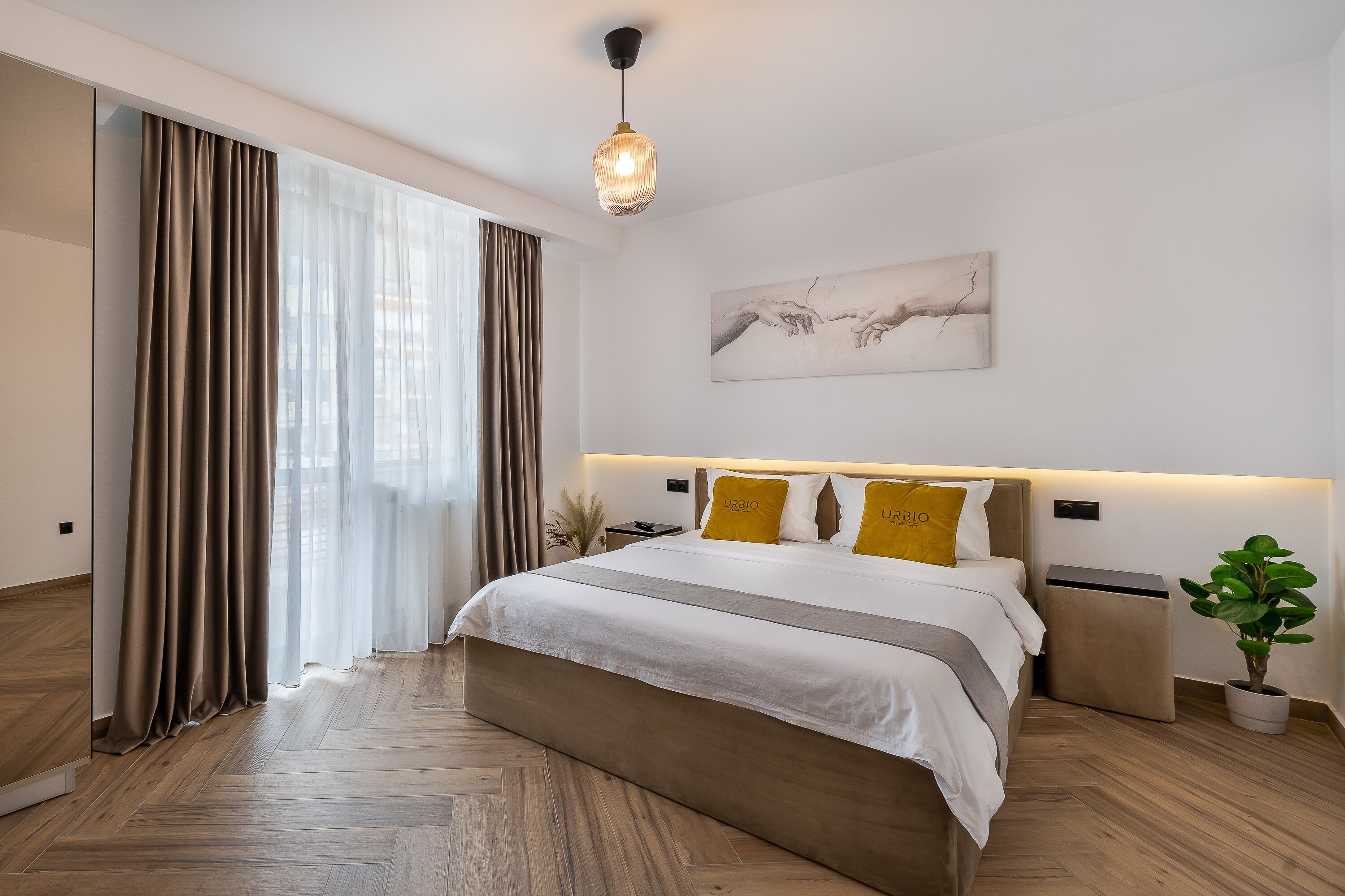 Moderni hotellihuone, jossa on suuri sänky, yöpöytä, pieni sivupöytä, ruukkukasvi ja taulu seinällä.