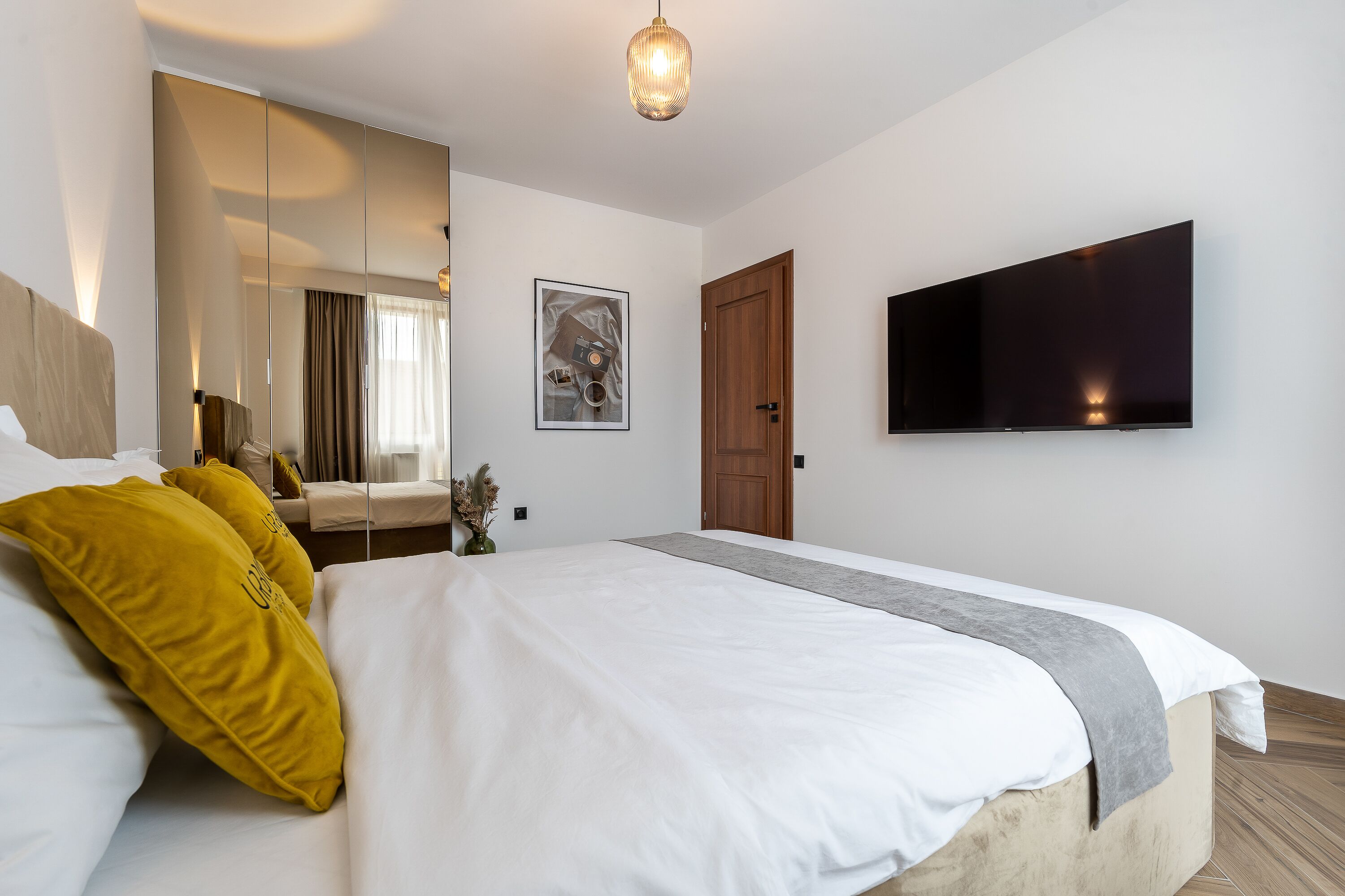 Een moderne hotelkamer met een groot bed, een flatscreen-televisie aan de muur, een houten deur en een spiegel die de kamer weerspiegelt.