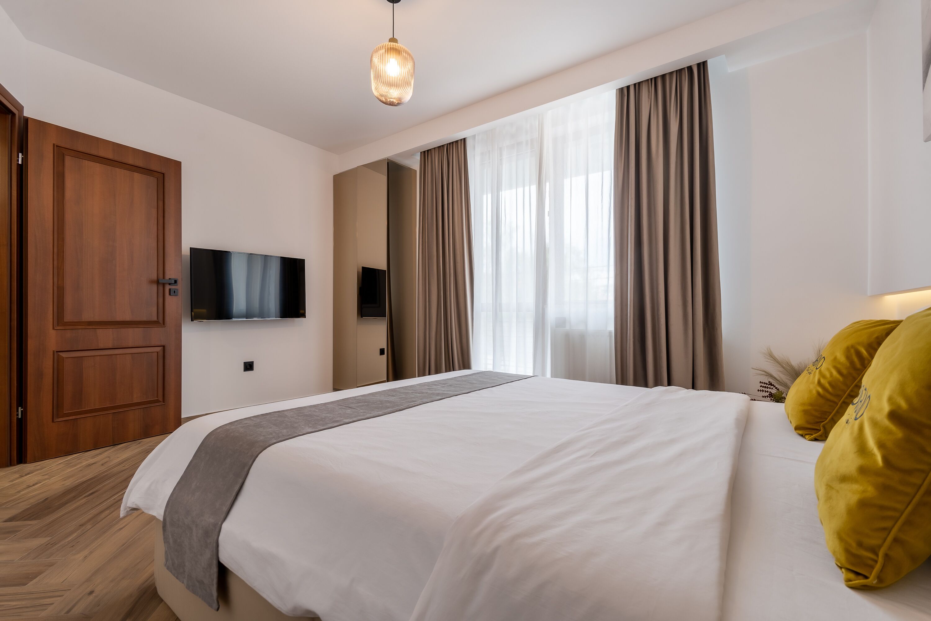 A hotel room with a large bed, a wooden door, a flat-screen TV mounted on the wall, and a window with curtains.