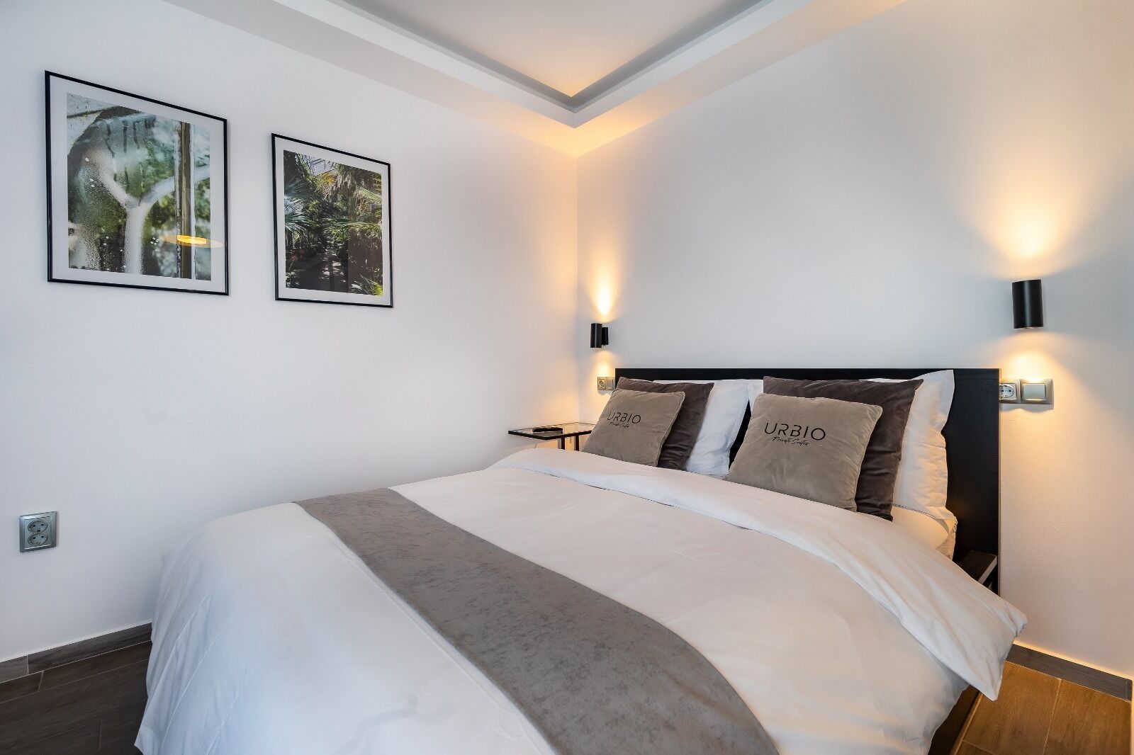 A modern hotel room with a large bed, two wall-mounted lamps, and two framed pictures on the wall.