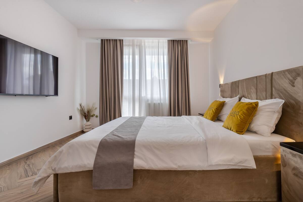A modern hotel room with a large bed, a flat-screen TV mounted on the wall, and a large window with curtains.