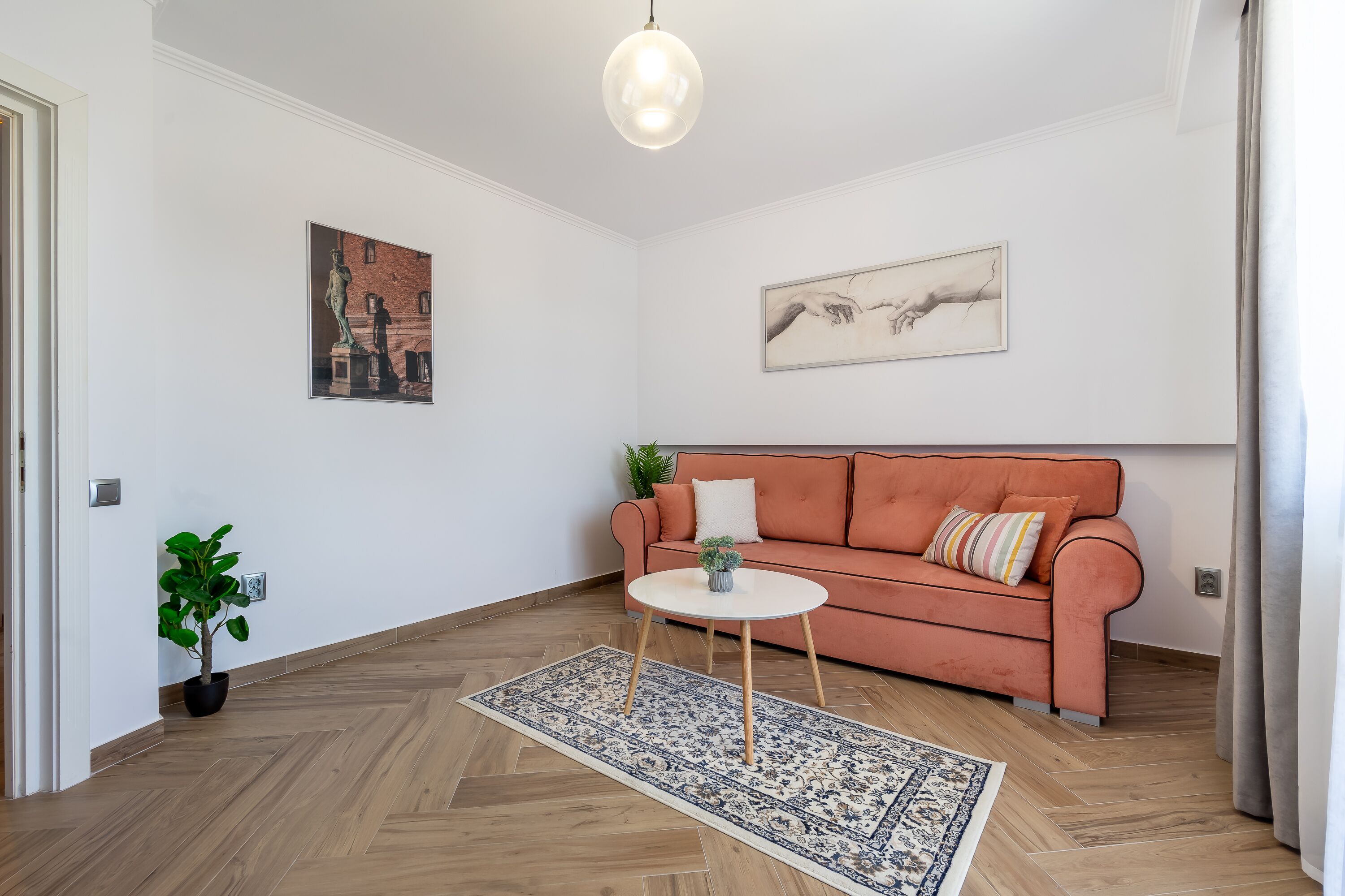 Een woonkamer met een visgraat parketvloer, een witte bank met kleurrijke kussens, een klein rond tafeltje en twee kunstwerken aan de muur.