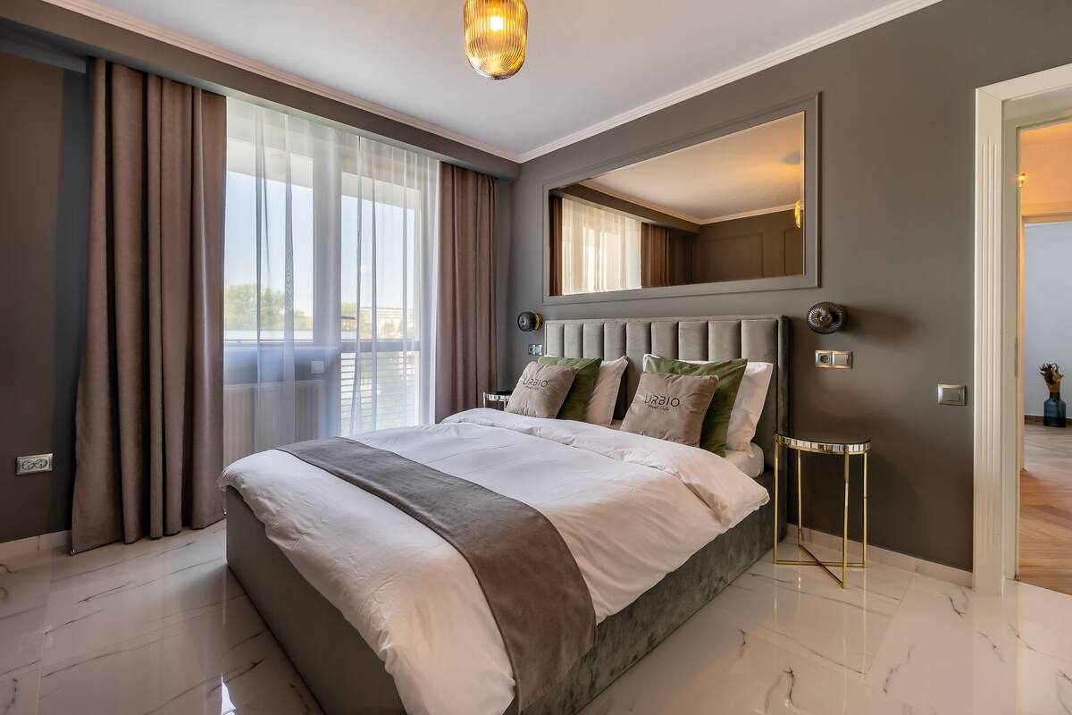 A modern bedroom with a large bed, a mirror above the bed, and a view of the outdoors through large windows.