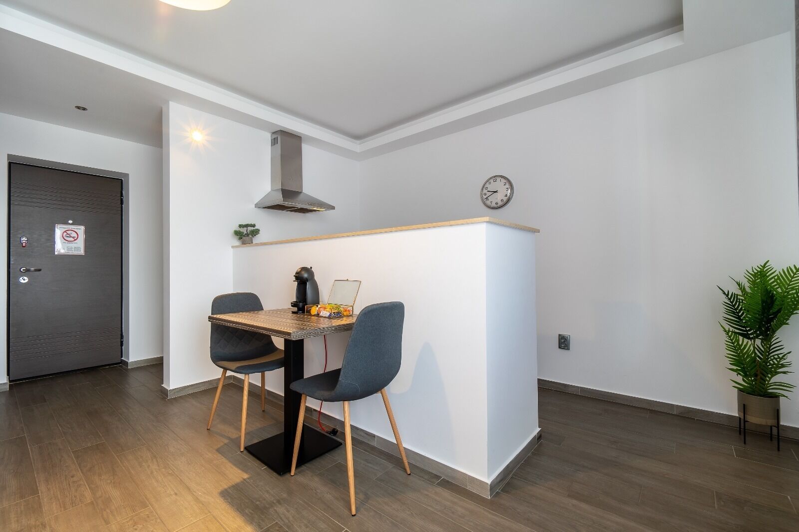 A small, modern kitchen with a dining table, chairs, a clock, and a plant.