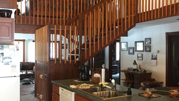 A spacious kitchen with a wooden staircase, a large island, and a dining area in the background. - Private 27 acres in peaceful Tapiola MI   Great home base to explore Western UP. (Chassell)