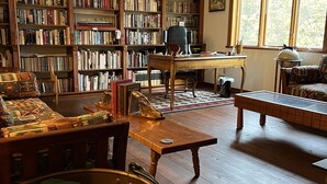 A cozy library with wooden bookshelves filled with books, a comfortable seating area with a patterned sofa and armchairs, and a large window providing natural light. - Private 27 acres in peaceful Tapiola MI   Great home base to explore Western UP. (Chassell)