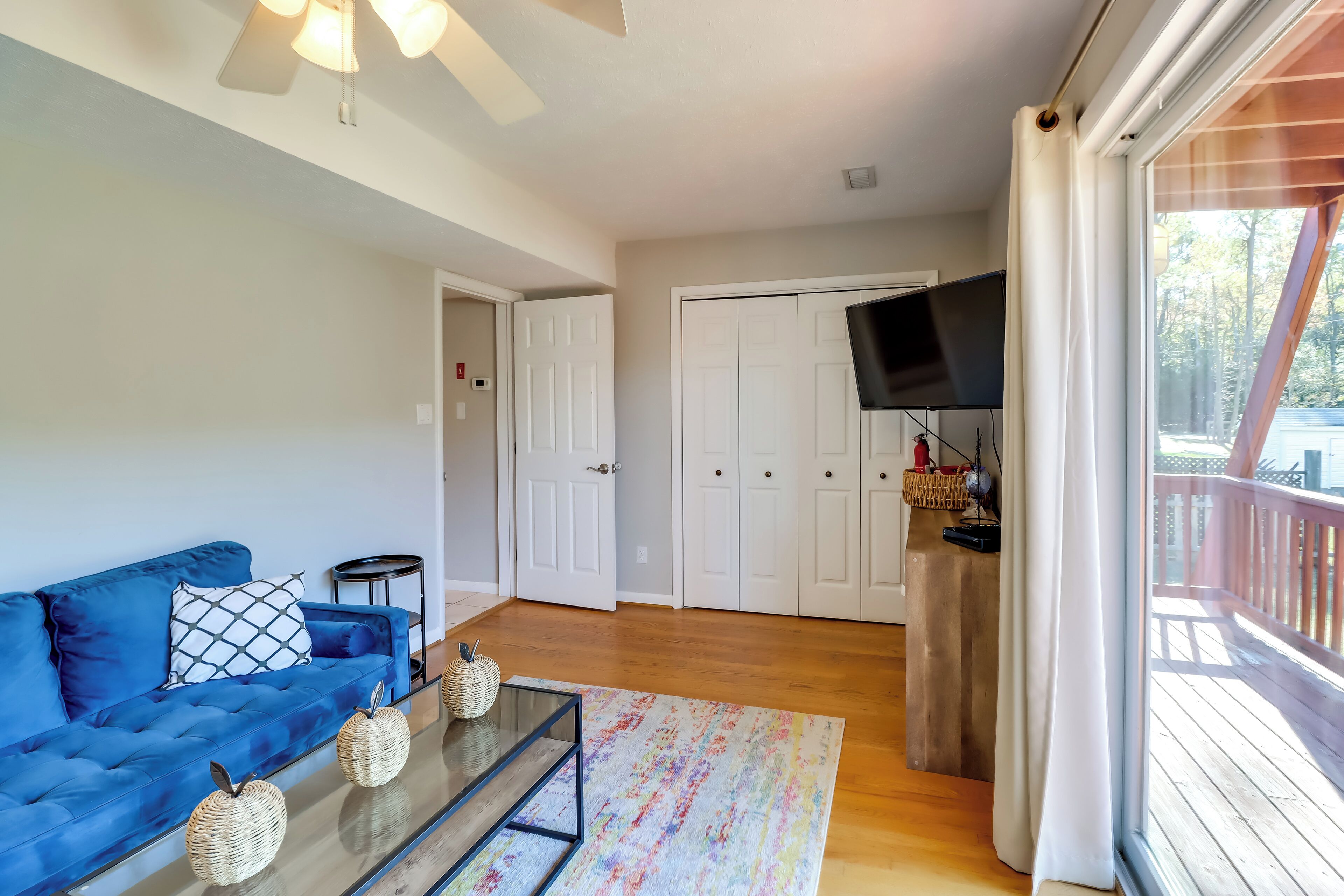 A living room with a blue sofa, a glass coffee table, and a colorful rug. There is a white door and a TV mounted on the wall.