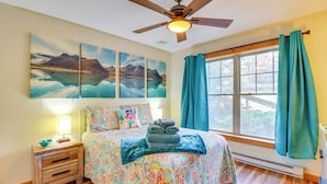 Quarto com cama, ventilador de teto, janela com cortinas e uma obra de arte na parede.