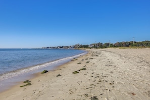A sandy beach with seaweed and footprints, a clear blue sky, and distant buildings. - Walk to Beach & Main St Hyannis: Cape Cod Home! (Hyannis)