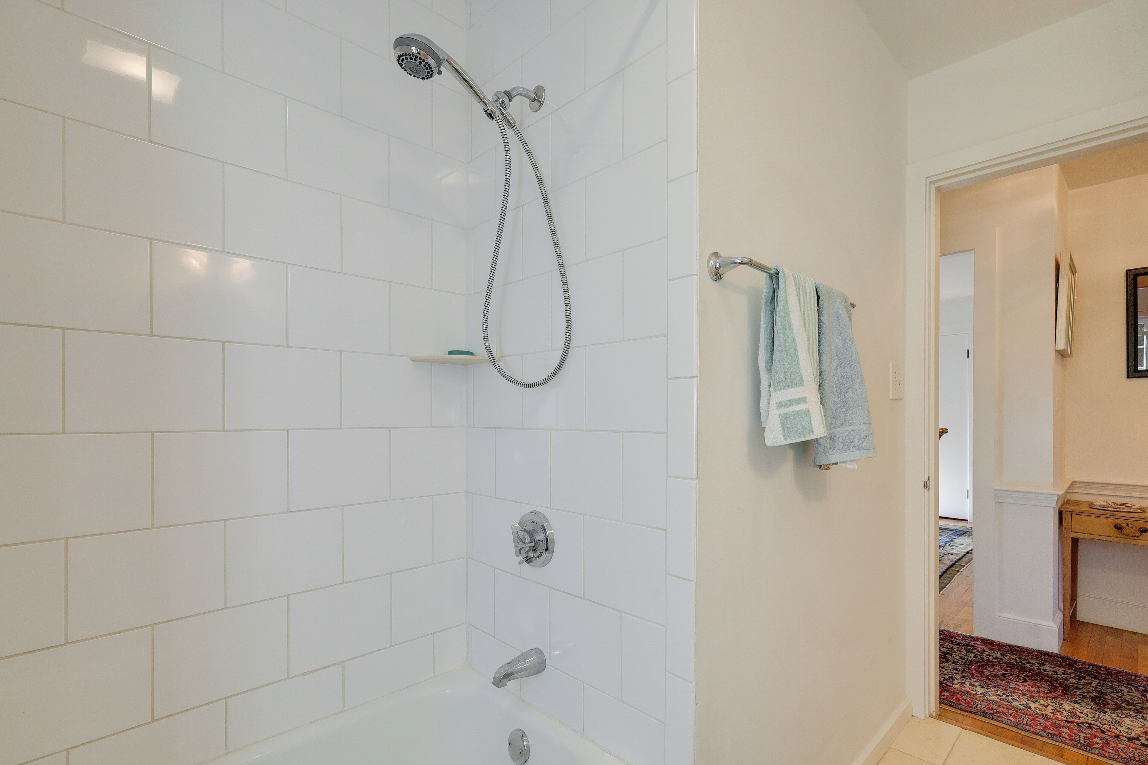 A modern bathroom with white tiles, a shower, a bathtub, and a visible hallway leading to another room.