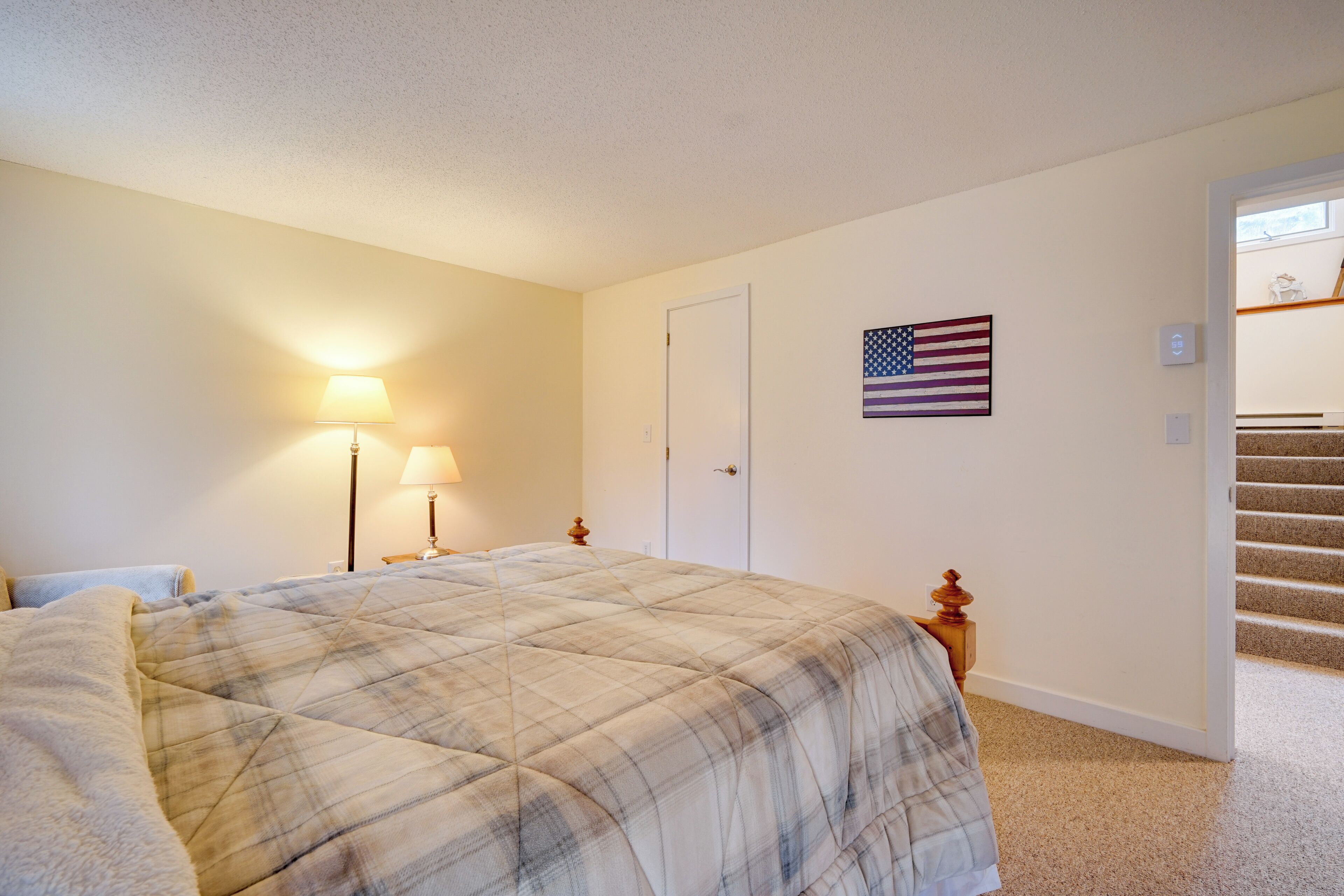 A bedroom with a plaid bedspread, a nightstand with a lamp, a door, and an American flag on the wall.