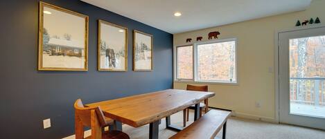 A dining area with a wooden table and benches, framed pictures on the wall, and a view through the window.