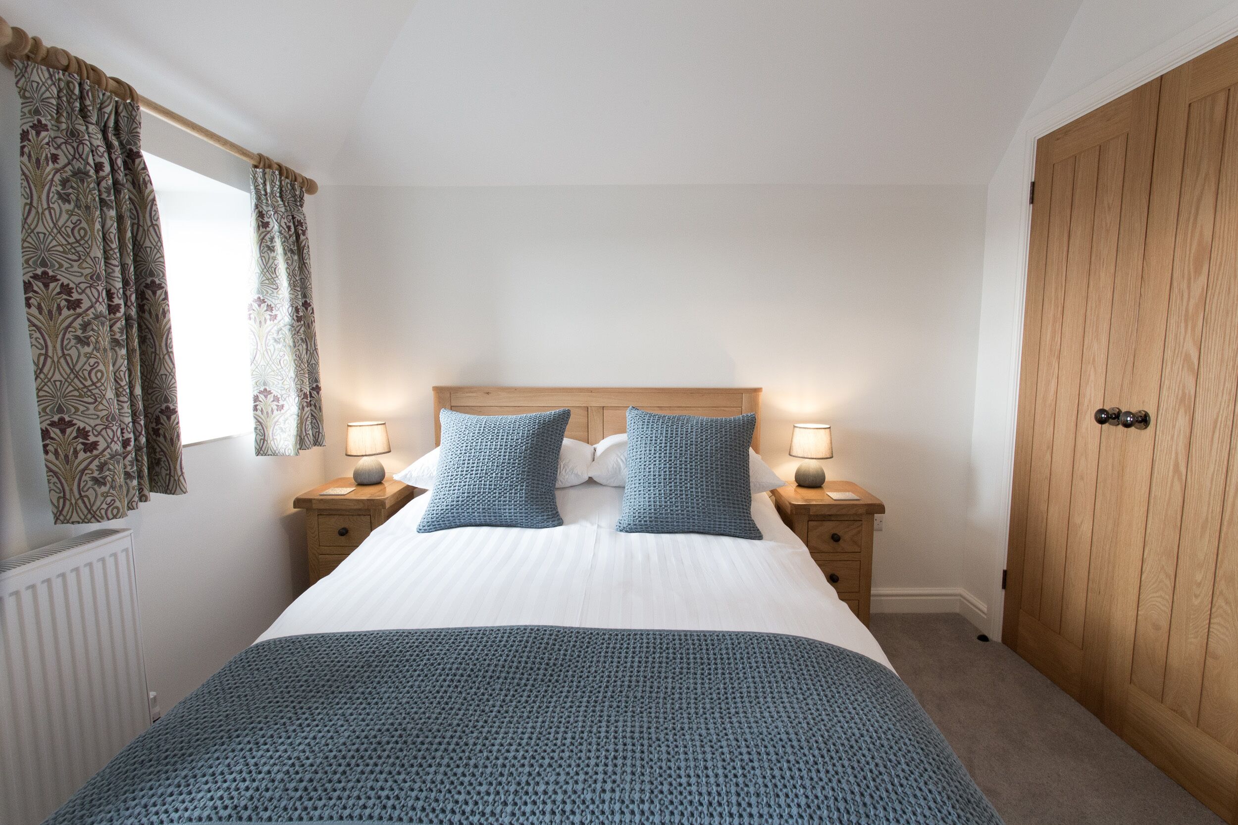 A bedroom with a large bed, two bedside tables with lamps, a wooden wardrobe, and patterned curtains.