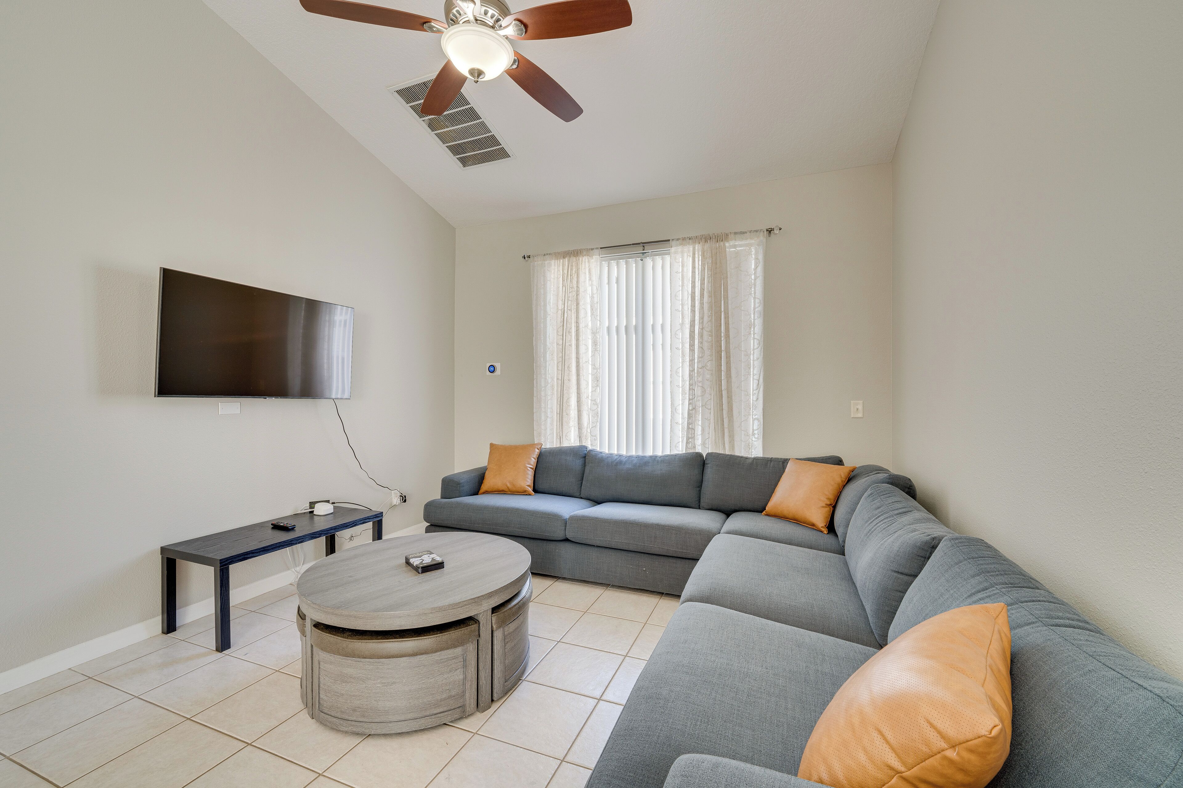 A living room with a grey sectional sofa, a round coffee table, a flat-screen TV mounted on the wall, and a ceiling fan.