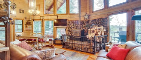 A cozy cabin interior with a stone fireplace, leather sofas, wooden floors, and large windows.