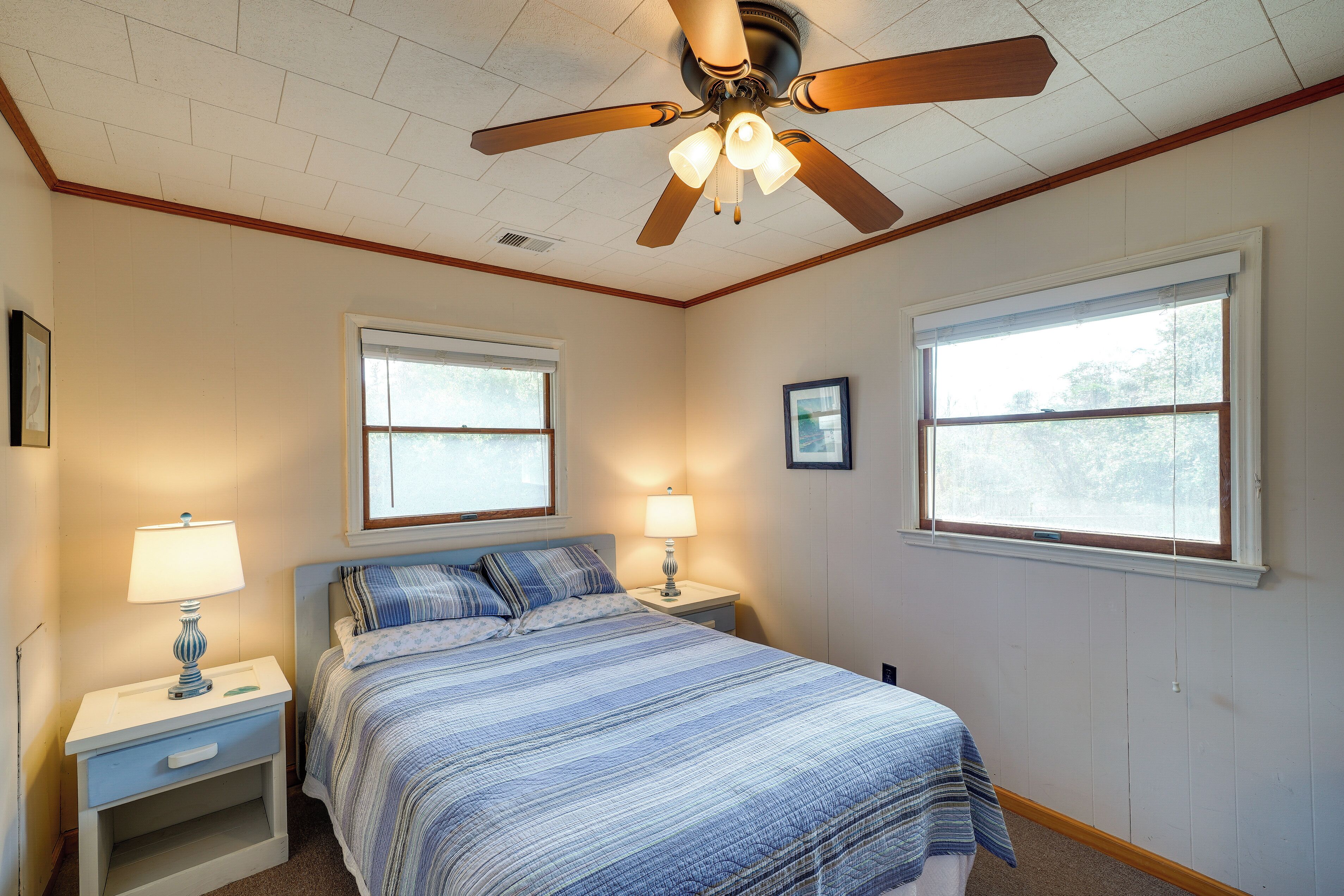 A bedroom with a bed, two bedside tables with lamps, a ceiling fan, and two windows.