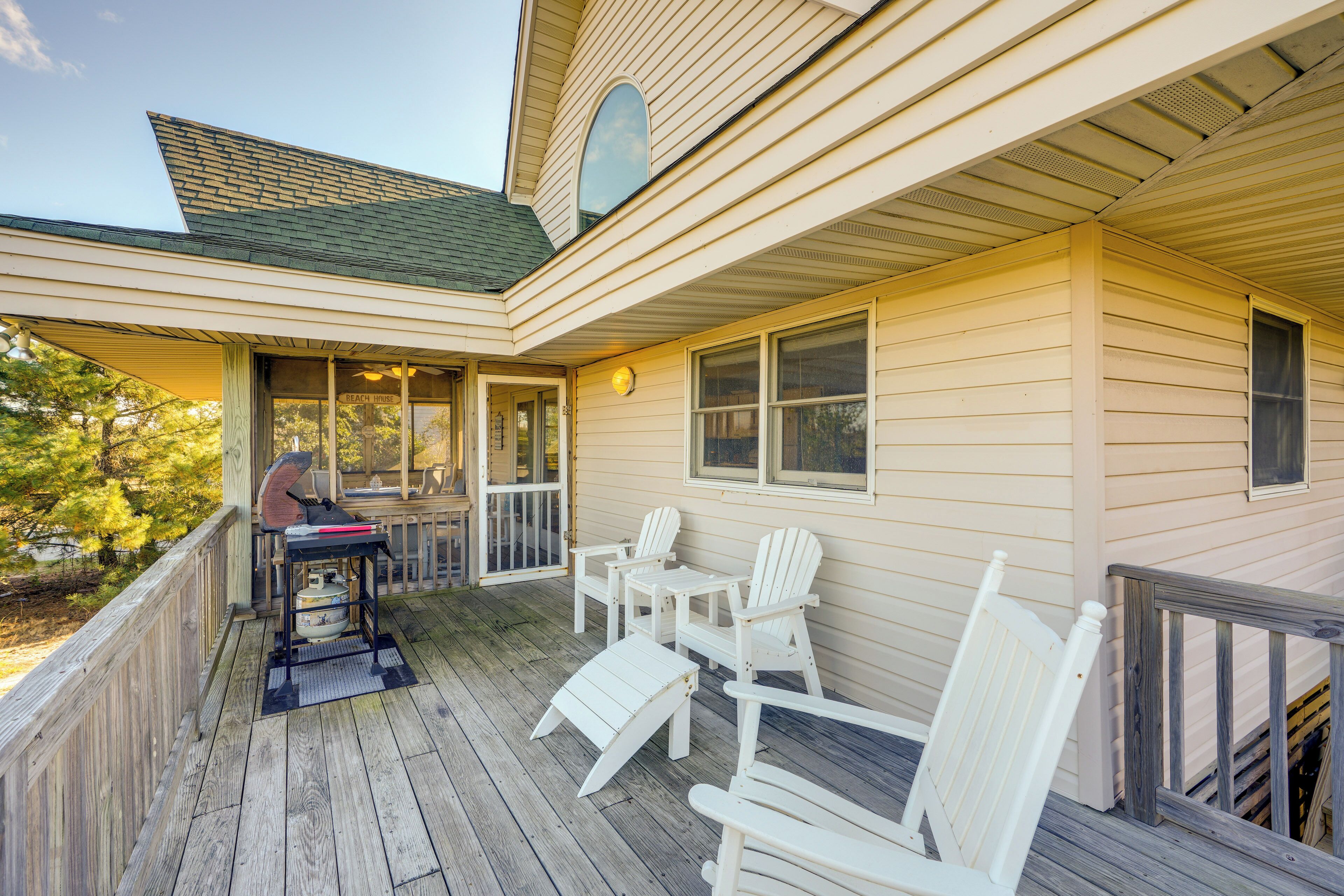 A wooden deck with white Adirondack chairs and a grill, overlooking a garden.