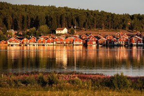 Un conjunto de casas rojas de madera frente a un lago tranquilo, con una colina boscosa al fondo.