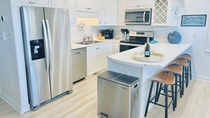 A modern kitchen with white cabinetry, stainless steel appliances, a central island with bar stools, and a decorative wall piece. - Oceanfront, Ocean Club F101, Linens included, 3Br 2ba, pool (Salter Path)
