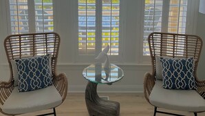 A room with two wicker chairs, a glass table with a white swan sculpture, and large windows with blinds. - Oceanfront, Ocean Club F101, Linens included, 3Br 2ba, pool (Salter Path)