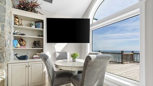 Un coin repas avec une table ronde, des chaises grises à revêtement rembourré, un buffet blanc agrémenté d’objets décoratifs et une grande fenêtre donnant sur la mer.