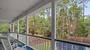 A covered porch with white railings, a rocking chair, and a view of a wooded area with a wooden fence and a blue pool. - Stay like a local in this pet-friendly home with private pool!  (Dauphin Island)