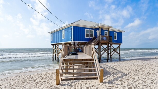 A wooden pier leading to a blue house on stilts over the beach.