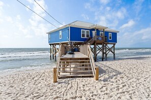 Un muelle de madera que conduce a una casa azul sobre pilotes frente a la playa.