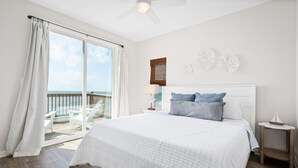 A bedroom with a large bed, a ceiling fan, a view of the ocean through a sliding door, and a wooden deck.