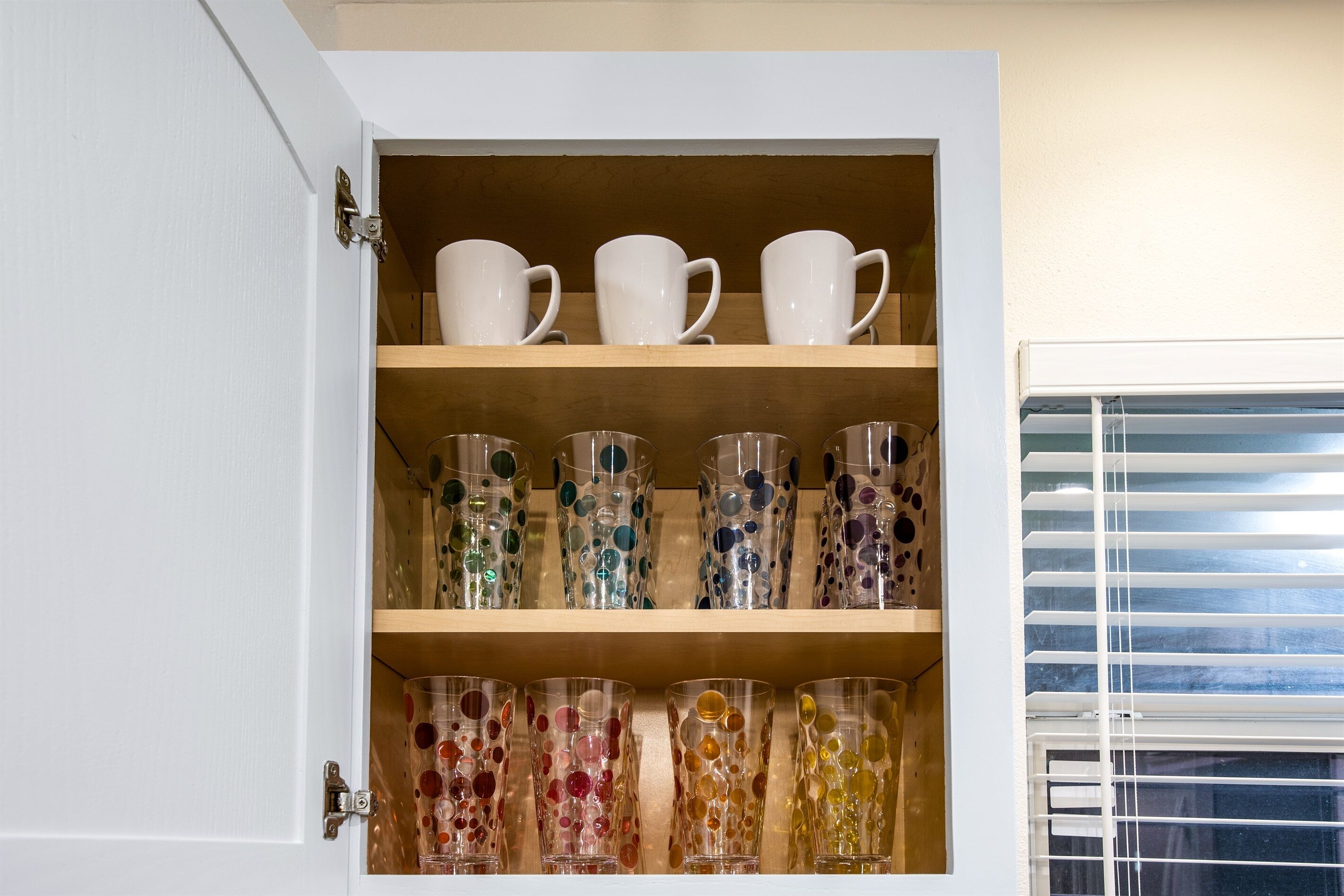 Un mueble con diferentes tipos de cristalería, incluyendo vasos transparentes y de colores, y tazas blancas en estantes.
