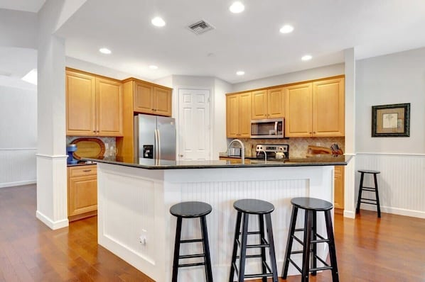 A kitchen with wooden cabinets, a central island with stools, a refrigerator, and a microwave.