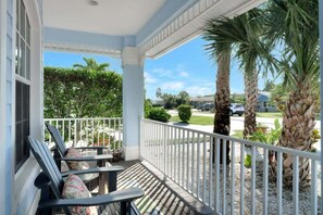 A covered porch with outdoor seating, palm trees, and a view of a residential street.