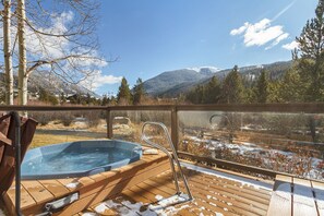 A wooden deck with a hot tub and metal railings overlooking a snowy landscape with mountains. - Cozy 2 Bedroom, walk to Peru Lift! (Keystone)