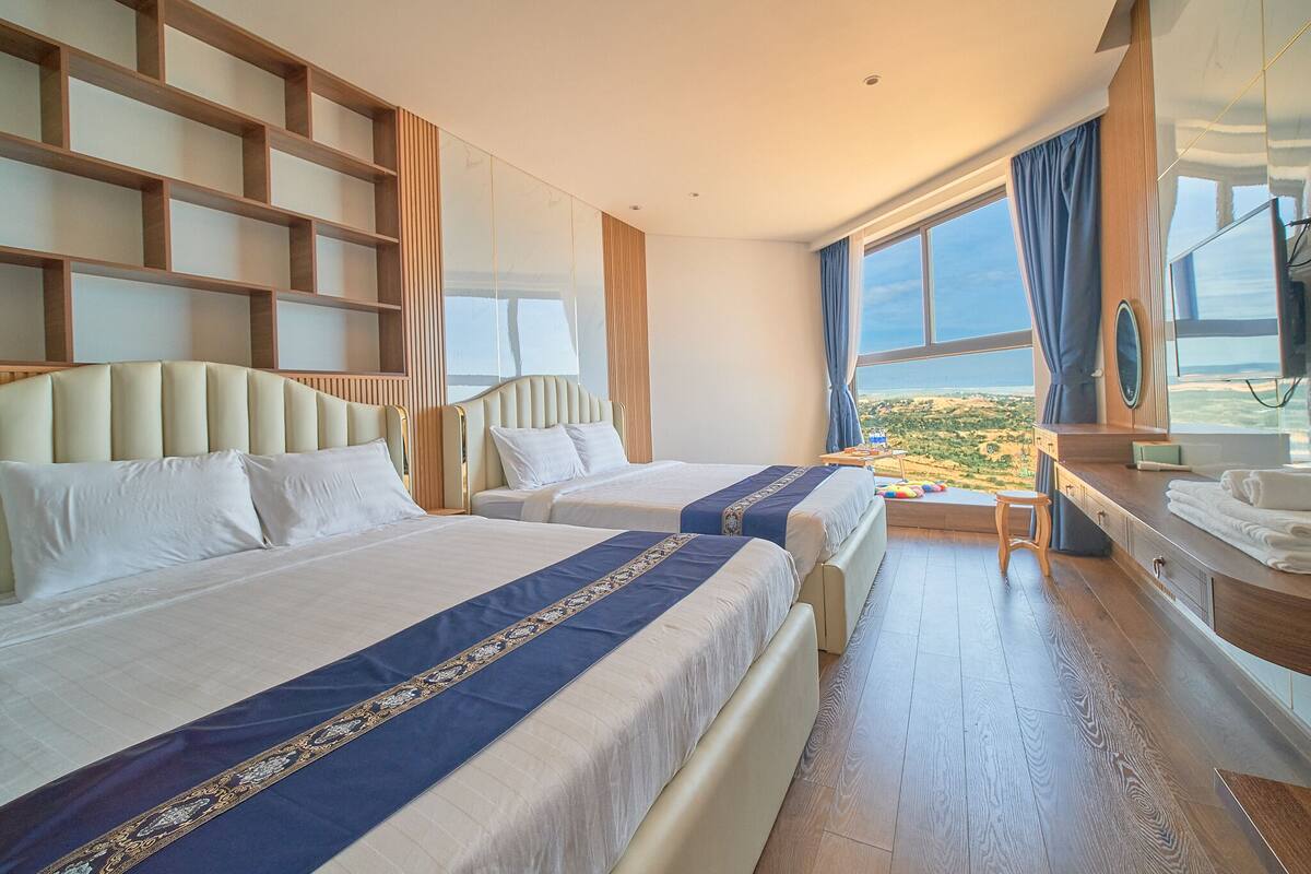 A hotel room with two beds, a large window with a view, a wooden desk, and a mirror.