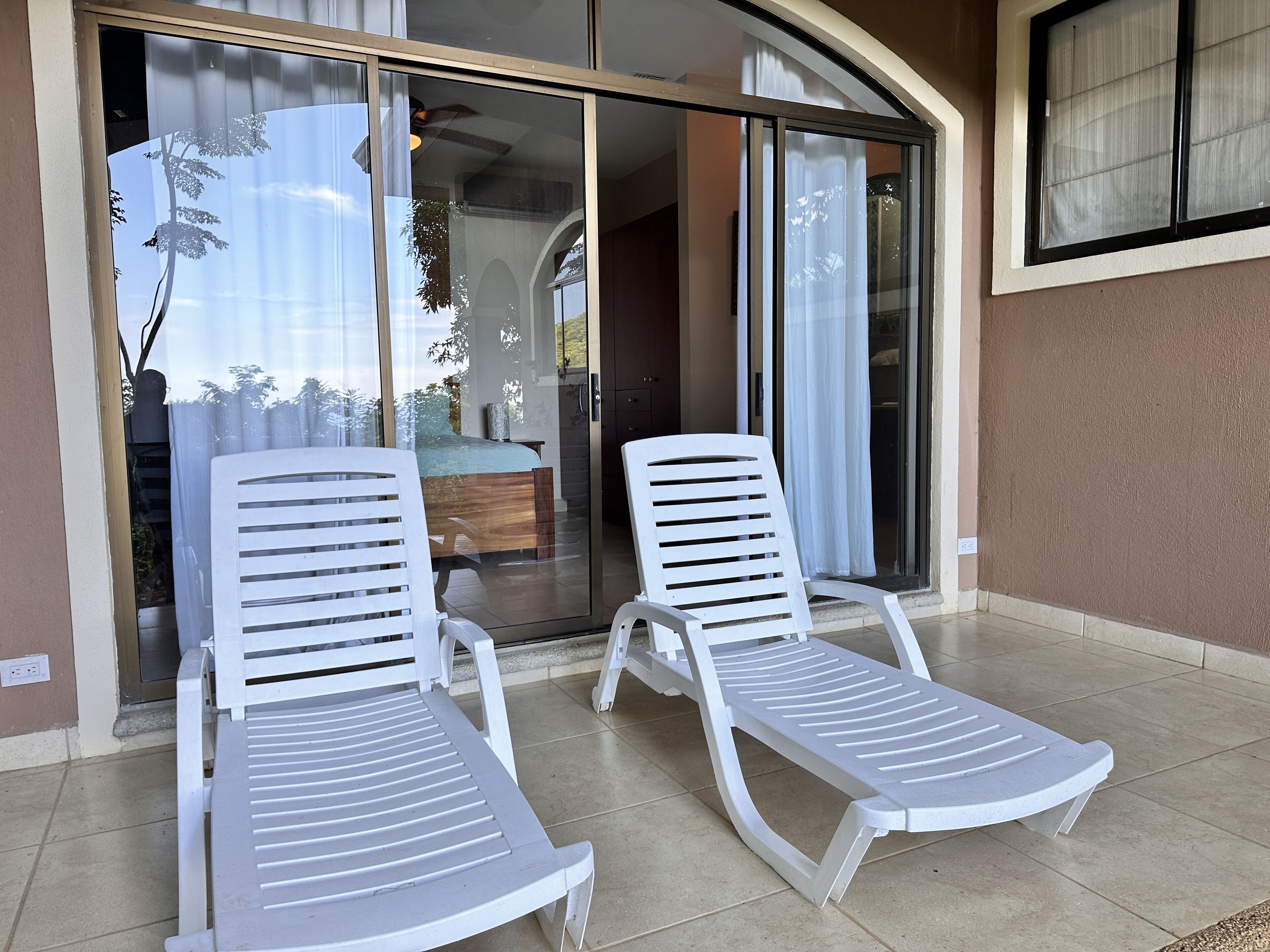 A balcony with two white lounge chairs, a glass door leading to a room, and a view of trees and a building in the background.