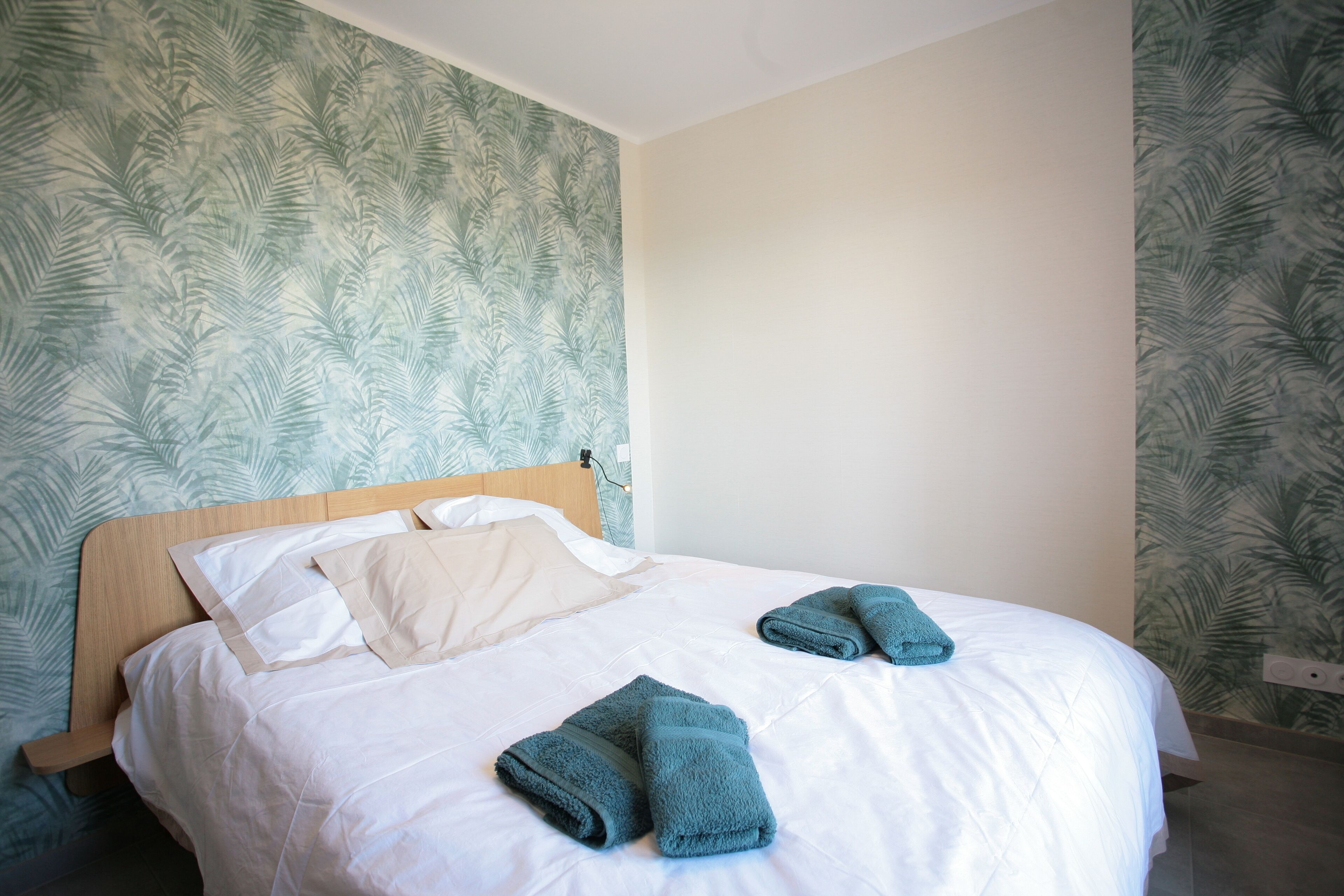 Un lit bien fait, avec du linge de lit blanc et deux serviettes pliées, une tête de lit en bois et un mur orné d’un motif végétal.