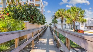 A wooden boardwalk leading to a building with balconies, surrounded by greenery and palm trees.