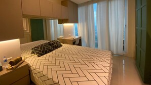 A modern hotel room with a large bed, a bedside table, a desk, and a view of the city through large windows. - Sky House BSD by Kakarama Room (Pagedangan)