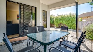 Een buitenpatio met een glazen tafel en stoelen, een schuifpui naar een balkon en een uitzicht op groen en een gebouw op de achtergrond.