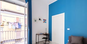 Ein modernes Wohnzimmer mit blauer Akzentwand, einem grauen Sofa und einem kleinen Schreibtisch mit Stuhl. Durch die Glastür ist ein Balkon mit Wäscheständer sichtbar.