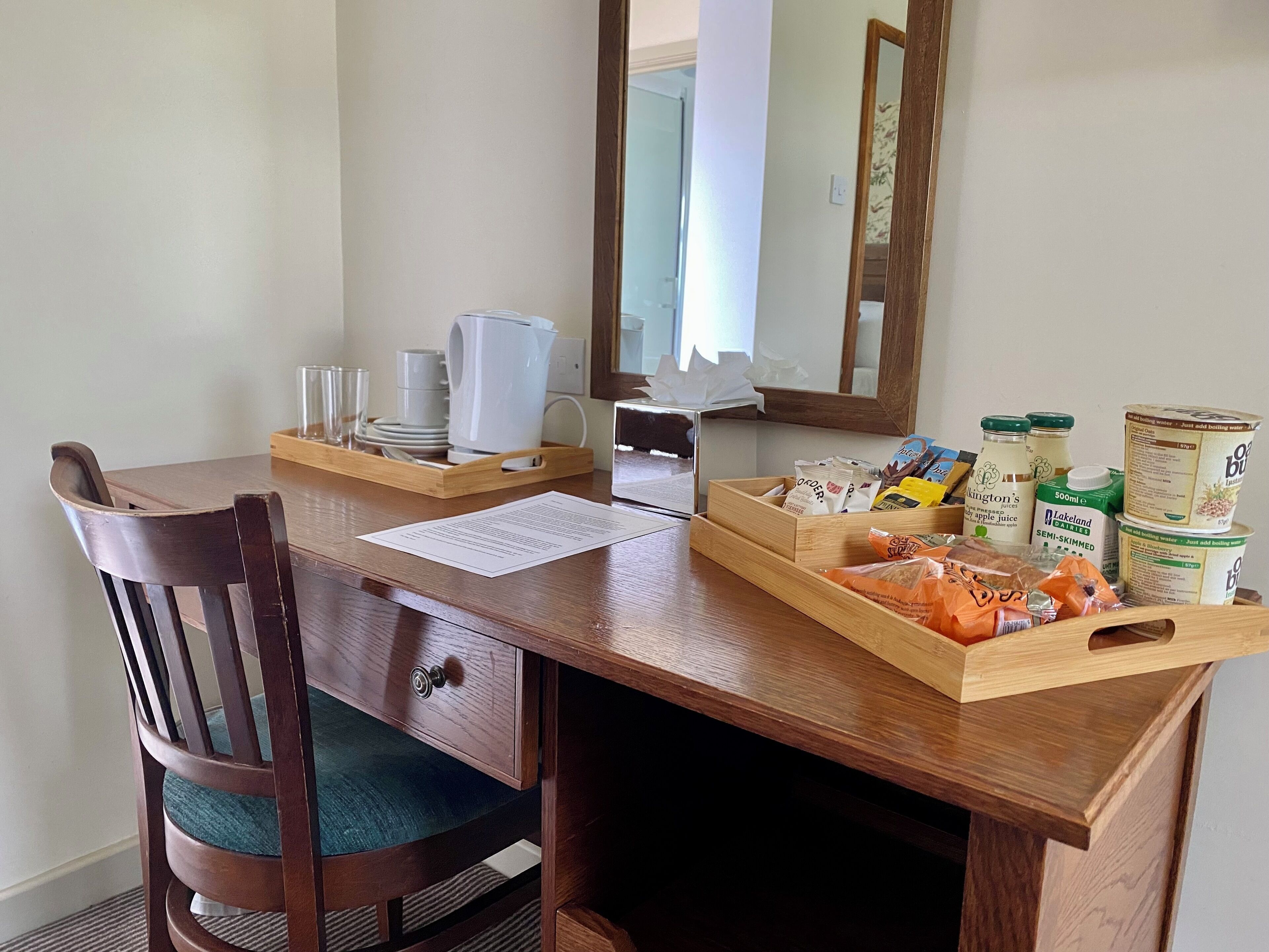 Ein Holztisch mit Stuhl, einem Spiegel und verschiedenen Gegenständen wie einem Tablett mit Snacks, einem Wasserkocher und einer Kaffeemaschine.