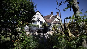 A white building with a steeply pitched roof and chimneys, surrounded by lush greenery and a patio area with outdoor furniture. - The Stag (Alcester)