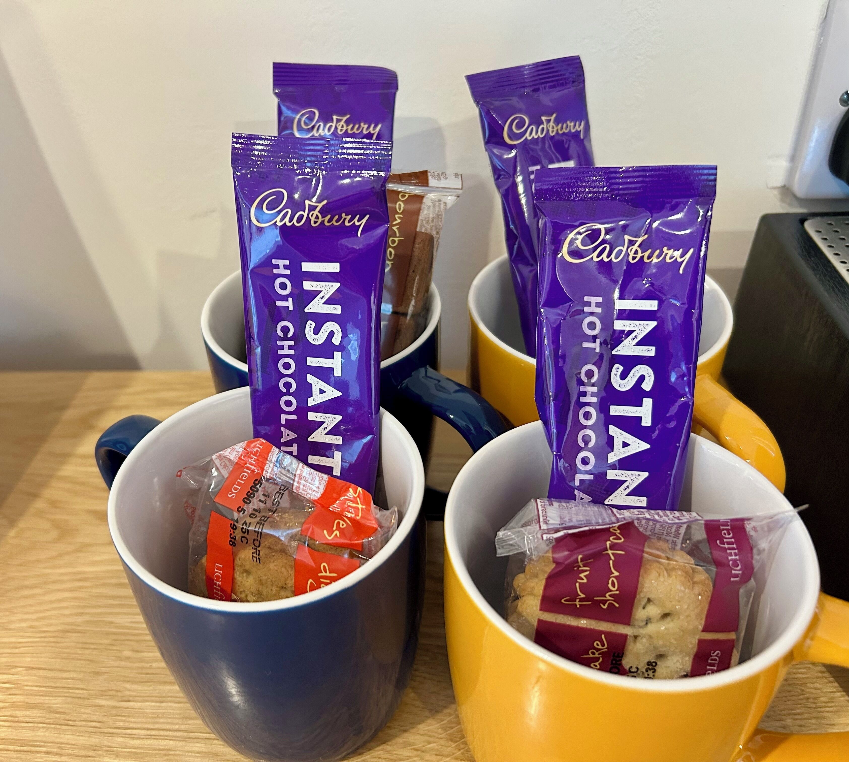 Tres tasses amb sobres de xocolata calent Cadbury i alguns biscotxes.