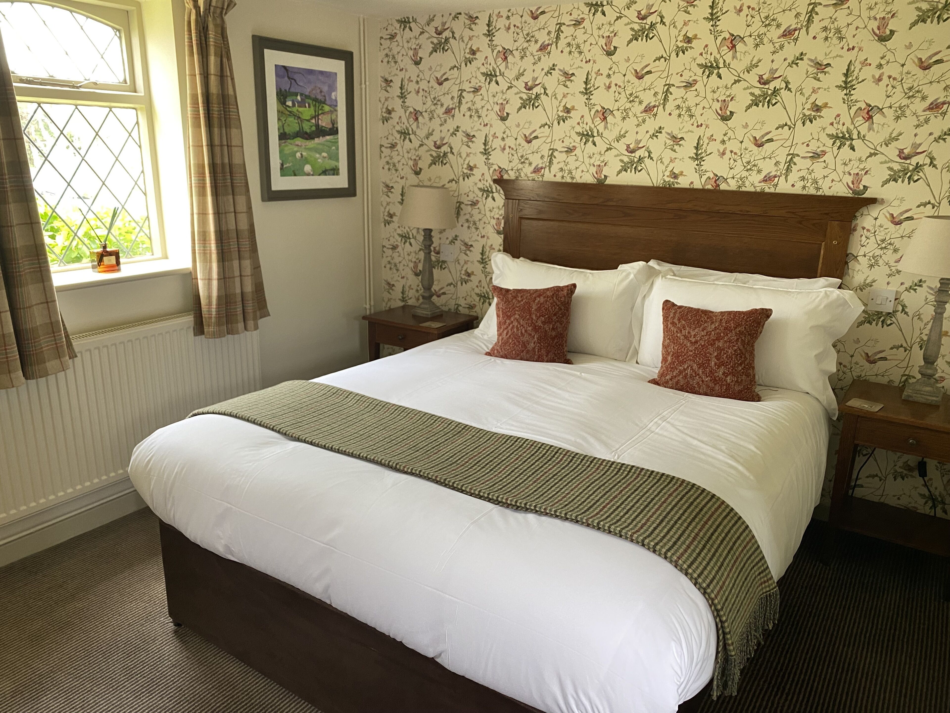 A neatly made bed with white linens, a brown headboard, and two decorative pillows. There is a window with curtains, a framed picture on the wall, and a small bedside table with a lamp.