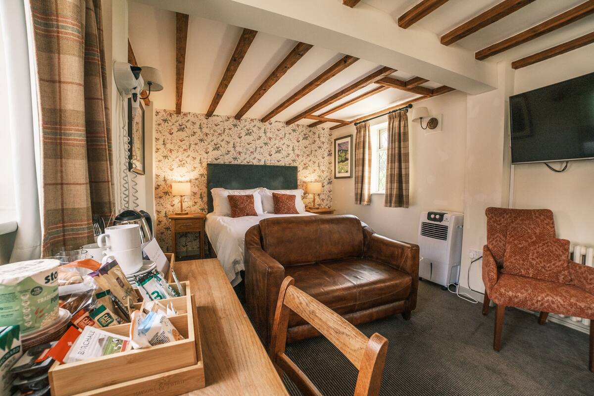 A cozy room with a bed, a wooden table with food items, a leather armchair, and a TV on the wall.