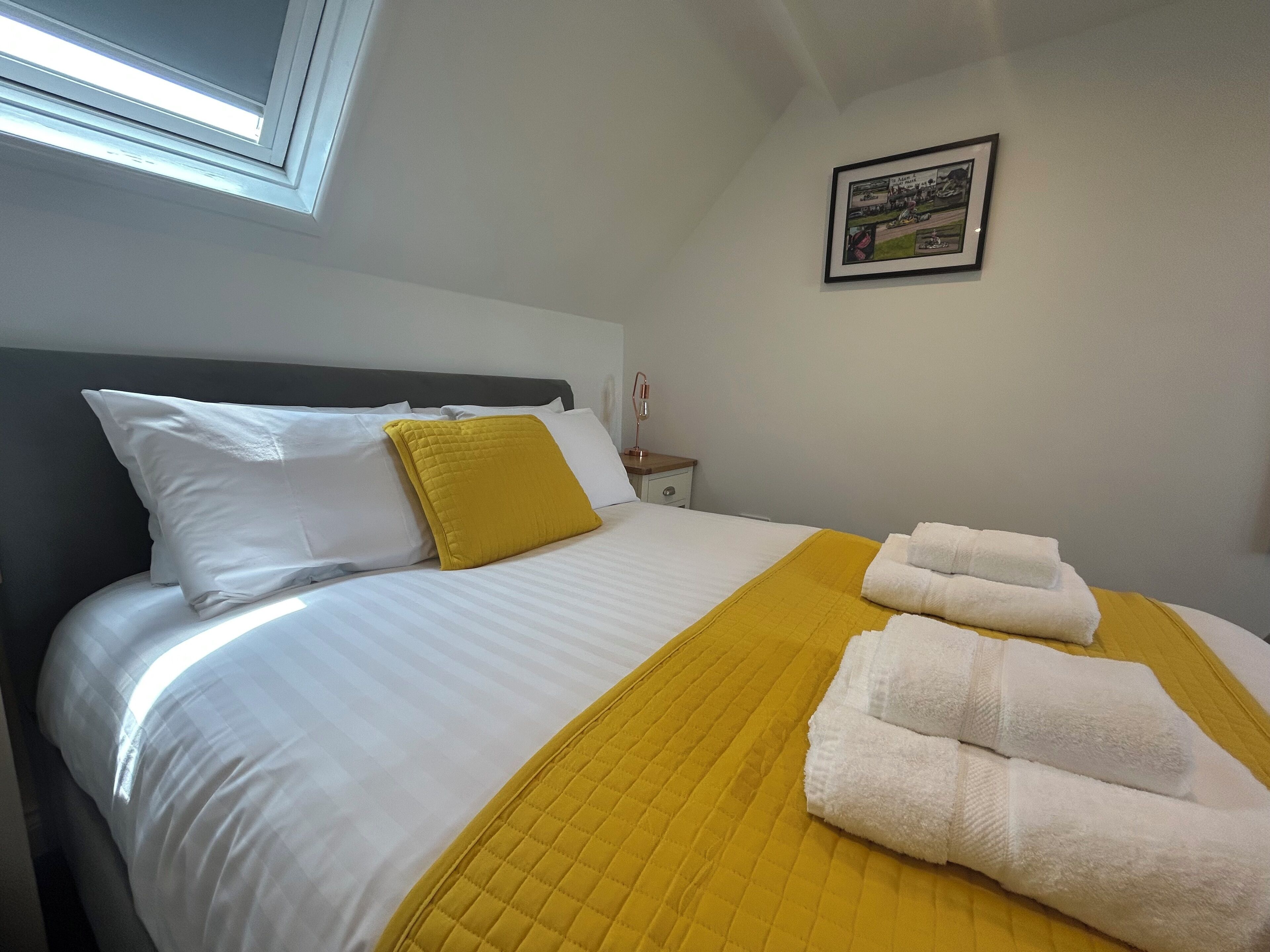A neatly made bed with white and yellow bedding, a framed picture on the wall, and a bedside table with a lamp.