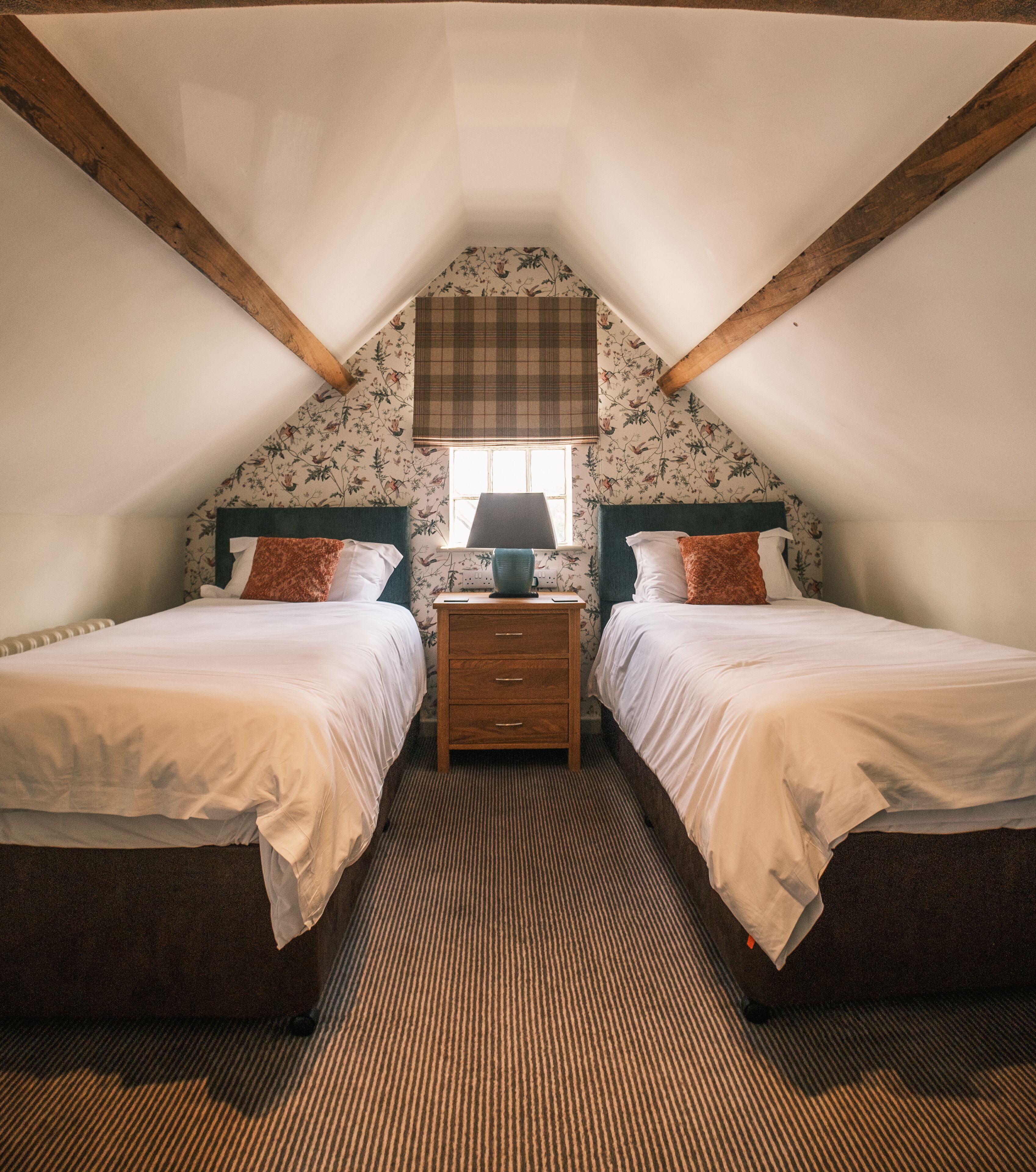 Une chambre avec deux lits, une table de chevet, une fenêtre avec un rideau à carreaux et une poutre en bois au plafond.