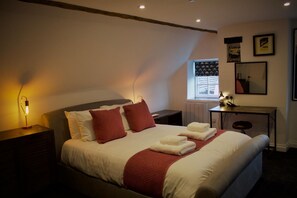 A neatly made bed with red pillows and white linens, a bedside table with a lamp, a desk with a chair, and a window with blinds.