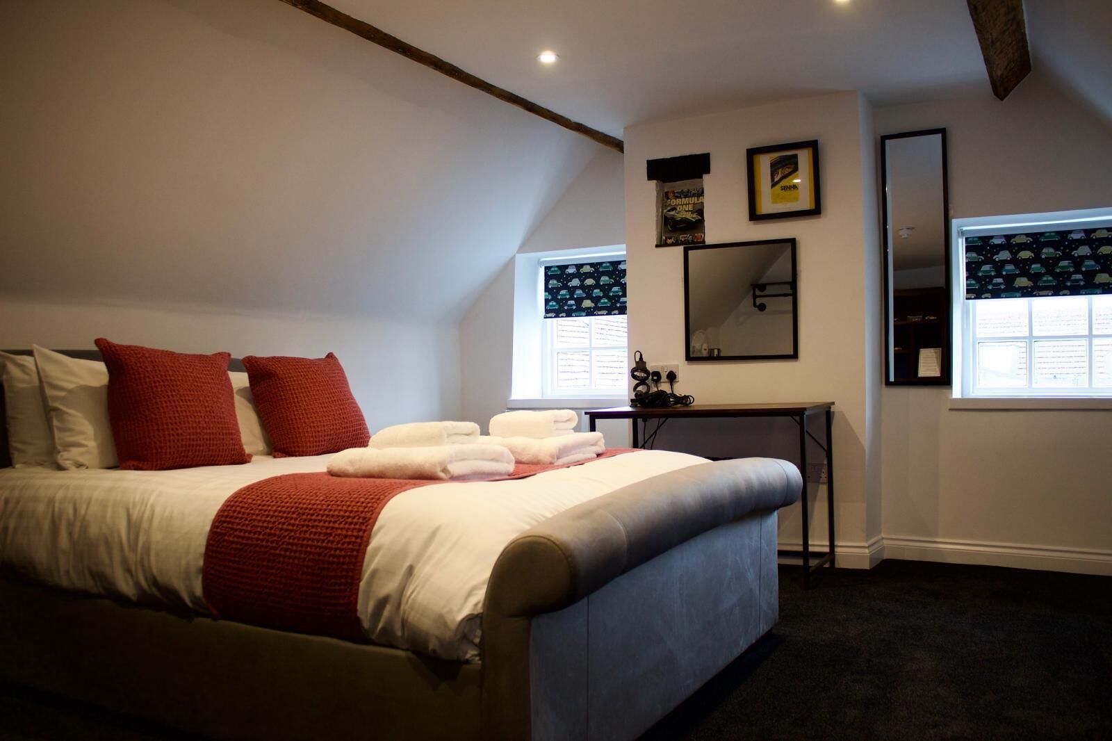 A bedroom with a bed, red pillows, a bedside table, a mirror, and a window with blinds.