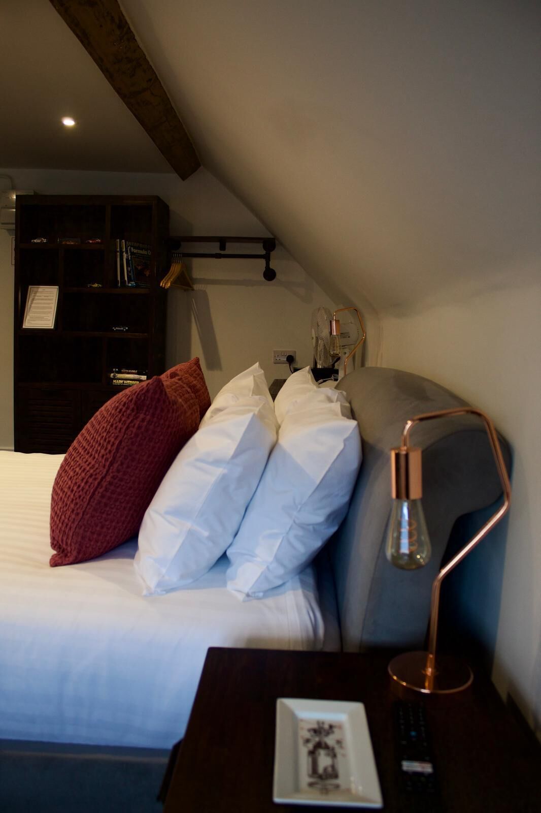 A bedroom with a bed, pillows, a bedside table with a lamp, a small table with a decorative item, and a bookshelf in the background.